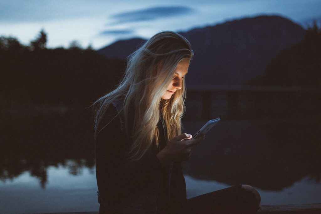 Woman sitting in front of a lake at night with a cell phone, the cell phone iluminates her face