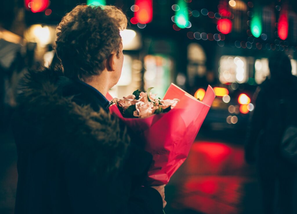 man waits at a bright lit up intersection carrying a bouquet of flowers