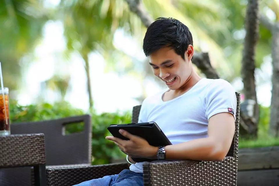 a man smiling while he looks at his tablet , sitting outside