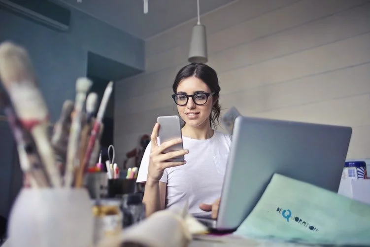 girl with glasses sitting in front of her computer checking her phone